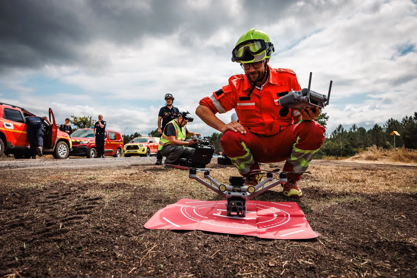 Un télépilote de la RODT accroupi près de son drone thermique posé, radiocommande en main, engins de secours à l'arrière-plan